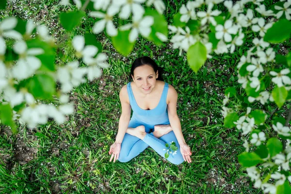 beautiful brunette woman in a sporty blue suit against a background of blossoming spring trees sitting on green grass and doing yoga relaxation, meditation concept