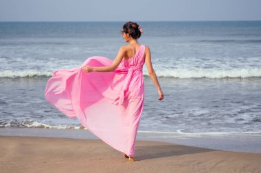 Genç ve güzel bir kadın plajda zarif, uzun, lüks bir elbiseyle dalgalarla poz veriyor. Tropik bir tatil beldesinde şık tatil kavramı