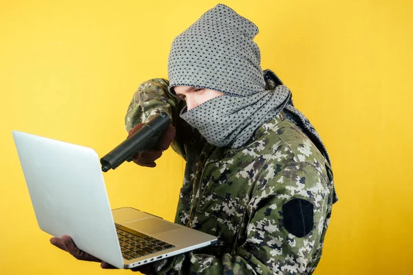 Portrait of a hacker man terroris in a camouflage and a mask is looking at the laptop