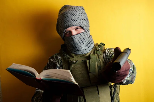 Ein Terrorist in Tarnkleidung und Maske hält ein rotes Buch und Waffen auf gelbem Hintergrund. das Konzept der Anonymität und des Terrorismus. — Stockfoto