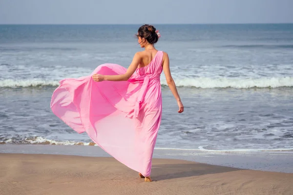Genç ve güzel bir kadın plajda zarif, uzun, lüks bir elbiseyle dalgalarla poz veriyor. Tropik bir tatil beldesinde şık tatil kavramı