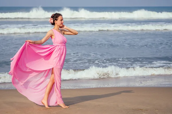 Genç ve güzel bir kadın plajda zarif, uzun, lüks bir elbiseyle dalgalarla poz veriyor. Tropik bir tatil beldesinde şık tatil kavramı