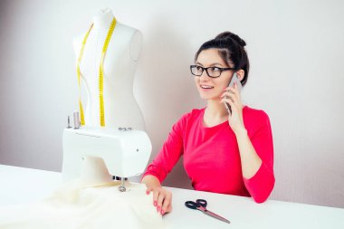 Gözlüklü, gülümseyen bir terzi kız telefonda dikiş makinesinden ve beyaz arka planda sarı bir mezura bulunan bir mankenden bahsediyor.