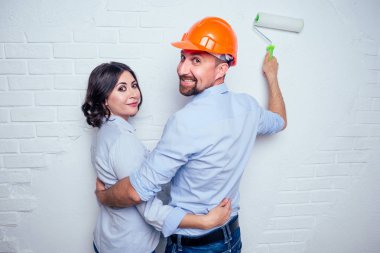 happy newlyweds family handsome man in the construction helmet hardhat and charming woman together painting white brick wall in house . concept of buying, repair and construction in house apartment