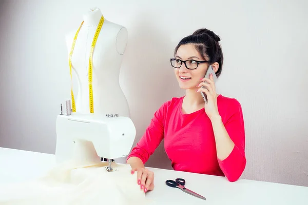 Costureira menina sorridente com óculos falando ao telefone sobre uma máquina de costura e um manequim com uma fita métrica amarela em um fundo branco — Fotografia de Stock