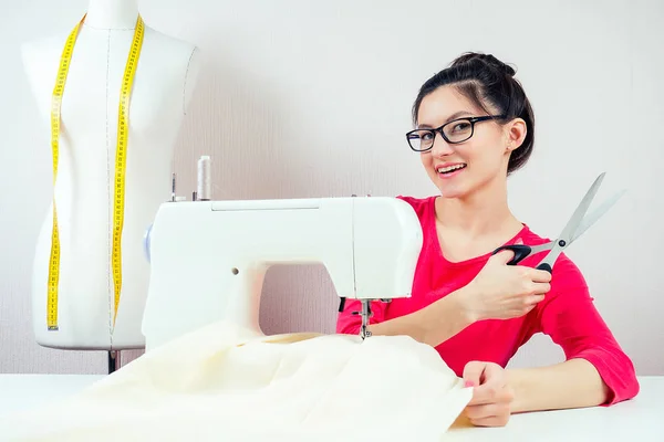 Улыбающаяся девушка-швея в очках, работающая с ножницами и швейной машинкой. На фоне манекена с желтой мерной лентой и белым фоном — стоковое фото