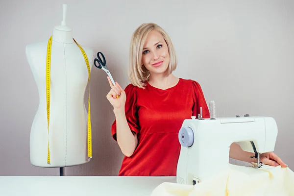 Costurera chica sonriente trabajando con tijeras y una máquina de coser. En el fondo del maniquí con la cinta métrica amarilla y el fondo blanco —  Fotos de Stock