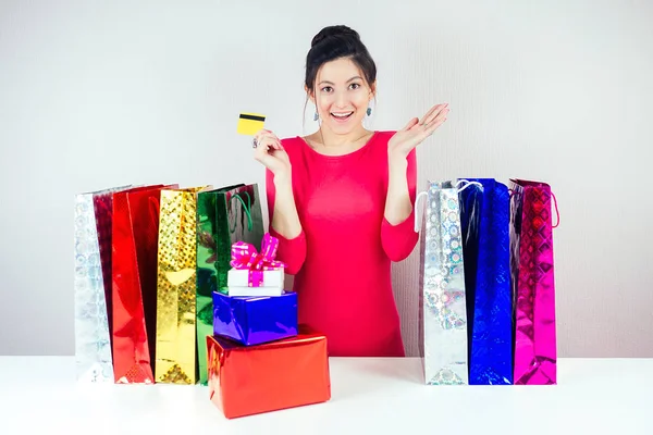 Attraktiv brunett kvinna i röd klänning med ett kreditkort förvånad och förvånad tittar på shoppingväskor och lådor med presenter. begreppet shopping för julklappar — Stockfoto