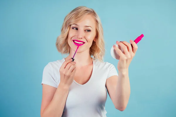 Attraente e sorridente donna bionda applica rossetto trucco rosa fucsia labbra in studio su uno sfondo blu — Foto Stock