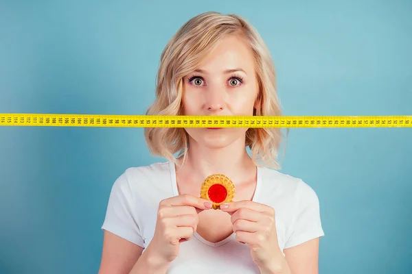 Atractiva mujer rubia triste con maquillaje y los labios de color fucsia sostienen una galleta de alto contenido calórico iwith cinta métrica n el estudio sobre un fondo azul. dieta y rechazo de los productos harineros — Foto de Stock