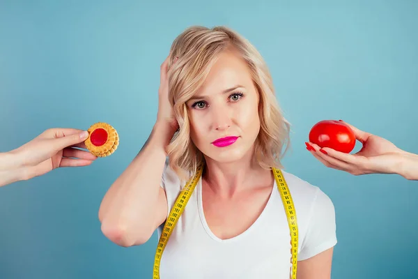 attractive blonde woman with make-up and lips color fuchsia makes a choice between high-calorie cookie biscuit and tomato with measuring tape in studio on a blue background. concept of healthy eating