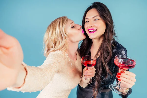 Due ragazze felici e attraenti bruna e donne bionde in abito da sera cocktail lucido paillettes stanno tenendo un bicchiere di vino e fare selfie al telefono a una festa in studio sfondo blu — Foto Stock