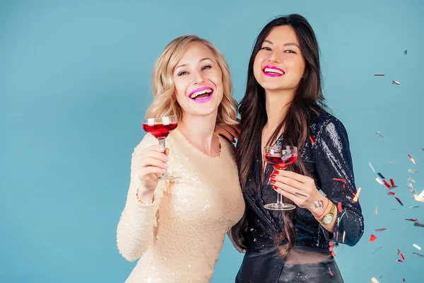 Due ragazze felici e attraenti bruna e donne bionde in abito da sera cocktail lucido paillettes stanno tenendo un bicchiere di vino a una festa in studio sfondo blu — Foto Stock