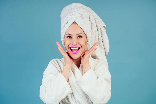 Atractiva y sonriente mujer sorprendida y sorprendida con un albornoz blanco de algodón y turbante de toalla en la cabeza en el estudio sobre un fondo azul. concepto de spa y frescura higiene femenina — Foto de Stock