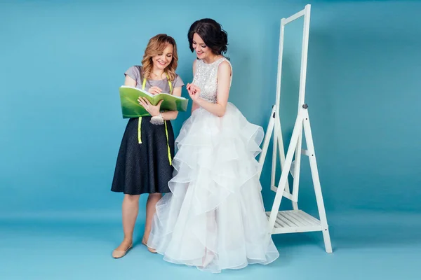 seamstress wedding consultant and bride discuss the details of wedding dress in the studio on a blue background . tailor dressmaker and customer prom in the dressing room next to the mirror