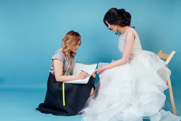 seamstress wedding consultant and bride discuss the details of party wedding dress in the studio on a blue background . tailor dressmaker and customer prom in the dressing room next to the mirror