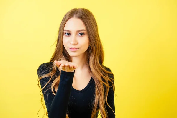 Porträtt av en ung attraktiv kvinna med långt hår skickar en luftkyss i studion på en gul bakgrund. konceptet Alla hjärtans dag och kärleksförklaring — Stockfoto