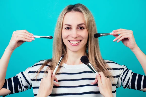 Retrato de una joven y hermosa multitarea visagiste mujer rubia maquillaje artista con maquillaje y peinado aplicar maquillaje un montón de manos y pinceles sobre un fondo azul en el estudio . — Foto de Stock