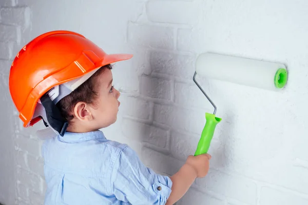 Affascinante carino bambino in casco hardhat dipinto parete con vernice di colore bianco. concetto di riparazioni casa appartamento — Foto Stock