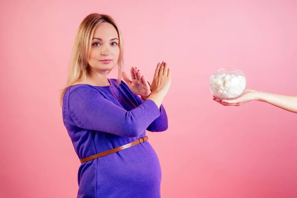 Beautiful blonde pregnant woman in a violet dress with big baby bump refuses sugar in the studio on a pink background . concept of healthy eating during pregnancy