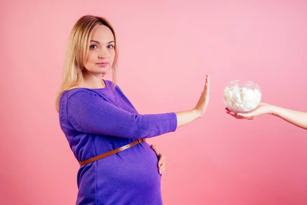 Stop eating sweets. Beautiful blonde pregnant woman in a violet dress with big baby bump refuses sugar in the studio on a pink background . concept of healthy eating during pregnancy