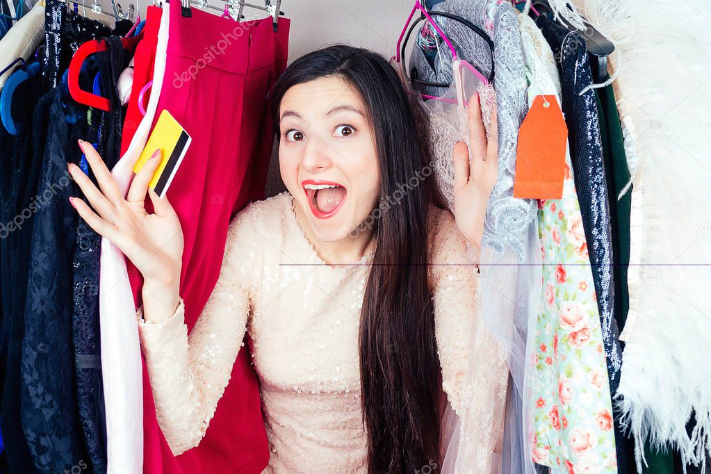 crazy brunette mad woman shopaholic surprised and amazed looks through the hangers wardrobe with clothes holding a plastic credit card in her hand . concept of seasonal sales and shopaholicism
