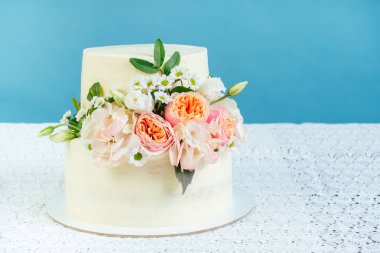 İştah açıcı, kremalı, iki katlı düğün pastası. Stüdyoda mavi arka planda dantel masa örtüsü olan bir masada taze çiçeklerle süslenmiş.