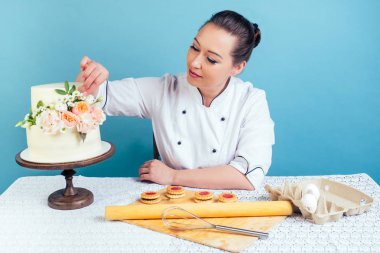Pastacı aşçı kadın kremalı, iki katlı düğün pastasını mavi arka plandaki stüdyoda taze çiçeklerle süslüyor. Tatil ve hazırlık etkinliği konsepti