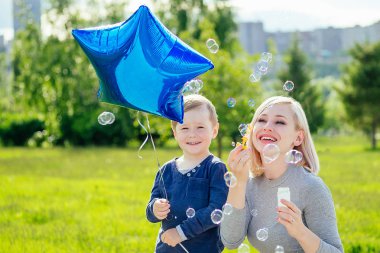 Güzel anne ve tatlı oğlu sabun köpüğü üflüyor ve yeşil çimen ve ağaçların arka planında hava mavisi helyum balonu tutuyor. Doğada aile tatili kavramı