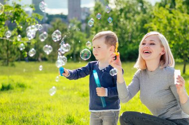 Güzel anne ve tatlı oğlu yeşil çimen ve ağaçların arka planında sabun köpükleri üflüyorlar.