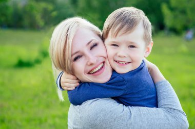 Çekici anne, parkta sevilen oğlunu yeşil çimenler ve ağaçların arkasında kucaklıyor.