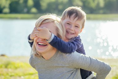 Güzel bir gülümseme anne ve tatlı oğlu çimenlerde oturup saklambaç oynuyorlar. Parkta, yeşil ağaçların ve nehir gölünün arka planında motosikletin yanında sarılıyorlar.