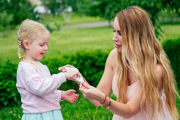 Little and sweet blonde girl in a cute gray dress together with toothy smile beautiful mother wipe hands with wet wipes in the park against the background of trees and greenery — Stock Photo, Image