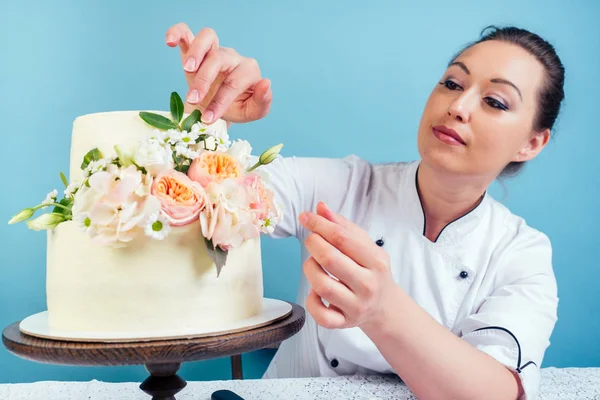 confectioner woman pastry-cook in confectioners jacket decorate appetizing creamy white two-tiered wedding cake with fresh flowers on a table with a lace tablecloth in studio on a blue background