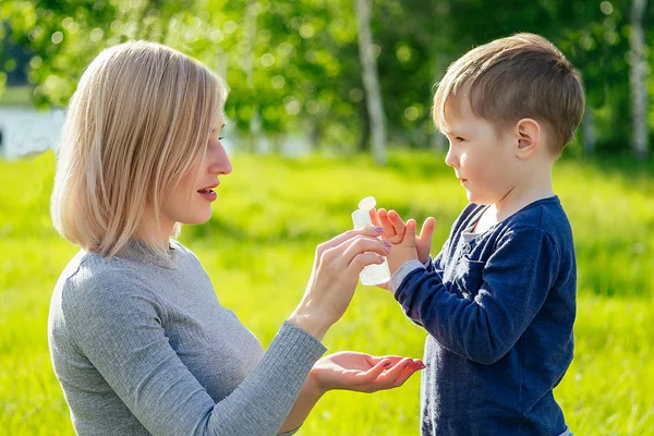 Güzel anne, oğullarına antiseptik jel sür. Parkta yeşil çimenler ve ağaçların üzerinde şirin bir bebek eli.