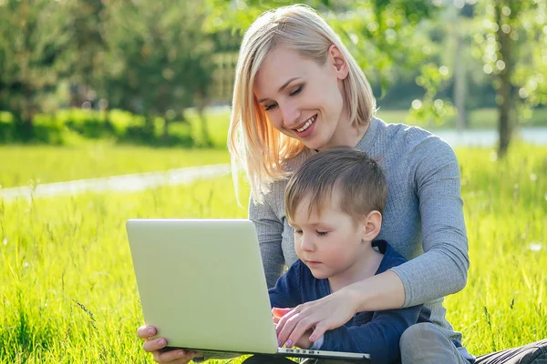 attractive mother female person and little boy watching cartoons on a laptop in the park on a background of green grass and trees
