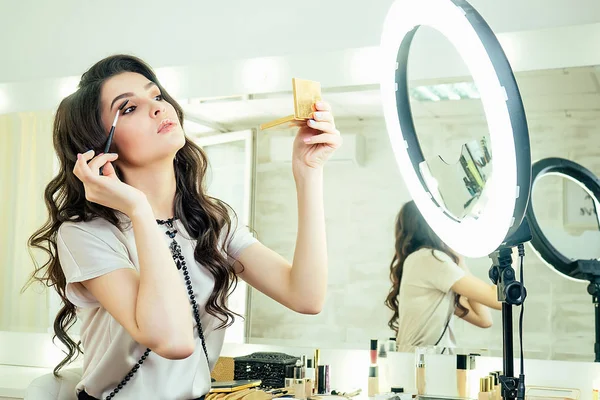 portrait of attractive and beautiful woman make-up artist visagiste sitting in front of a mirror and applies eyeshadow cosmetics make-up on face in salon studio. concept of self-care
