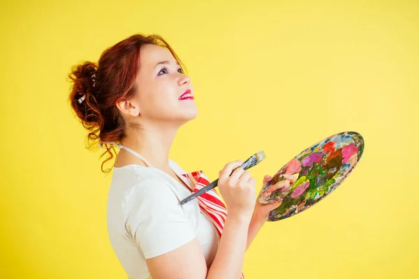 Portrait of a beautiful artist in an apron holding a palette and brush on a yellow background in the studio . inspiration and muse idea Royalty Free Stock Photos