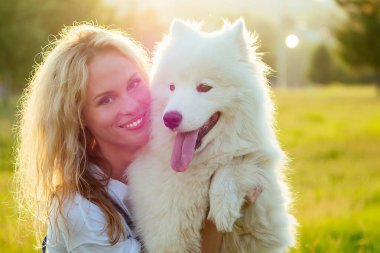 Güzel ve çekici kıvırcık saçlı, kot pantolon giymiş, sırıtan dişlek kadın camda oturuyor ve yaz parkındaki günbatımı ışınlarında tüylü beyaz bir köpeği öpüyor.