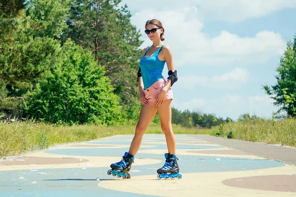 attractive young athletic slim brunette sexy woman in short pink shorts and blue top with protection elbow pads and knee pads posing on roller skates in the park