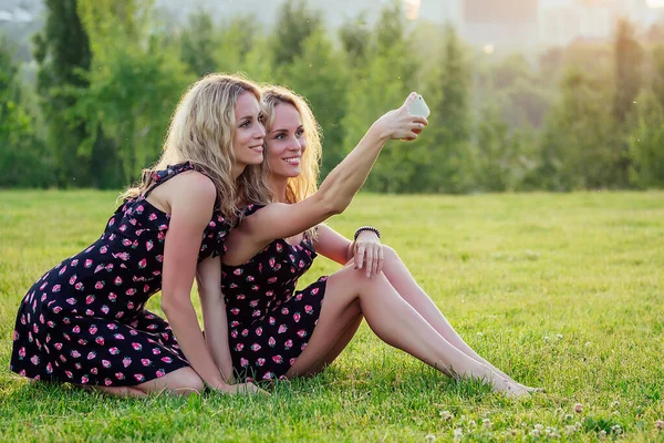 Zwei lustige Schwestern Zwillinge schöne lockige Blondine glücklich junge zahme Lächeln Frau in stilvollem Kleid haben Spaß und machen Selfie am Telefon im Sommerpark Sonnenuntergang Strahlen Feld Hintergrund — Stockfoto