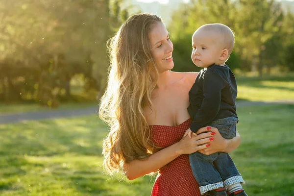 Zorgzame en mooie langharige vrouw in cocktail rode jurk houdt de baby jongen zoon in haar armen in het park in de zomer .love en zorg concept — Stockfoto