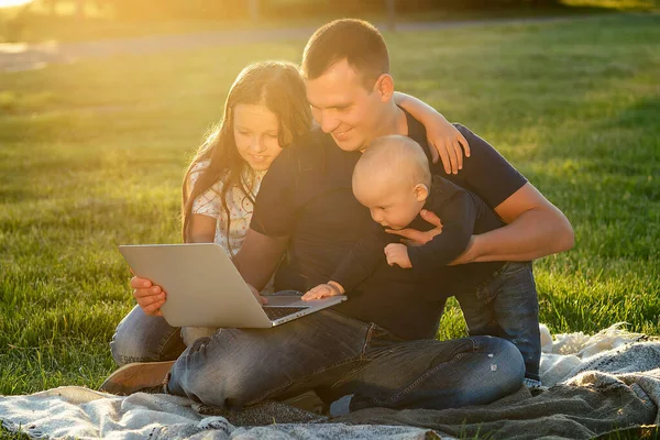 Niño pequeño junto con su padre y una hermosa hija jugar y desarrollar mirando en el ordenador portátil en una manta en el parque en verano — Foto de Stock