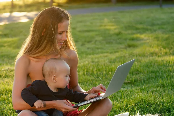 Idea de licencia de maternidad. encantadora y atractiva hermosa mujer con un hijo bebé que trabaja al aire libre en el parque de verano con el ordenador portátil sentado en la hierba césped verdes — Foto de Stock