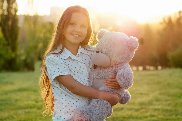 Bonito de cabelos longos bronzeado menina toothy sorriso segurando um cinza ursinho brinquedo em mãos no parque árvores fundo — Fotografia de Stock