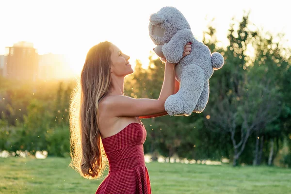 Schattig langharig gebruinde vrouw raadselachtige glimlach met een grijze teddybeer speelgoed in handen in het park bomen achtergrond — Stockfoto