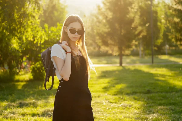Giovane adolescente con lunghi capelli biondi in uniforme scolastica vestito nero con zaino nel parco al tramonto — Foto Stock