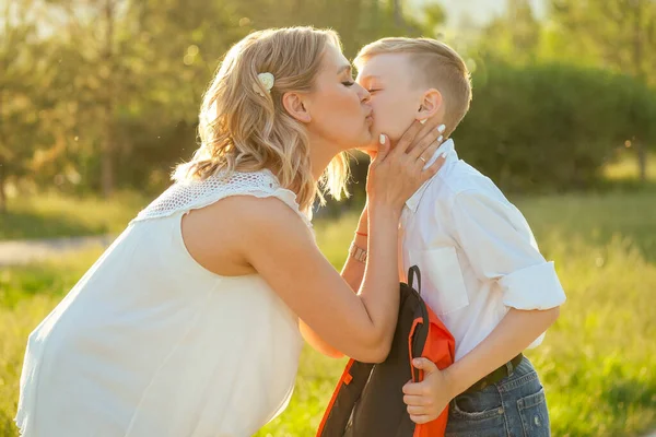mother kissing son before going to school . stylish and young schoolboy in a white shirt and jeans with a backpack in the park