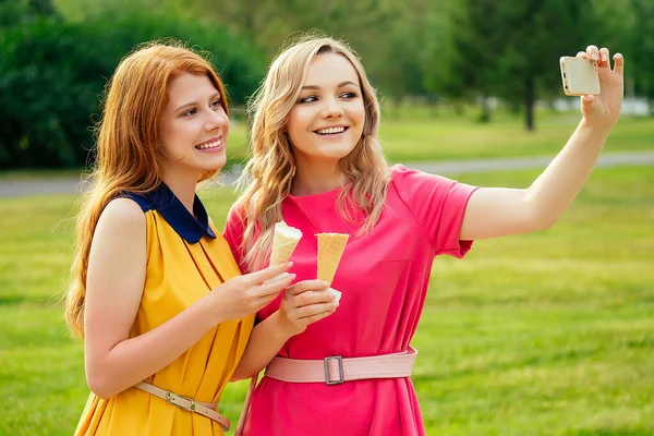 Zwei Freundinnen schöne junge Ingwerrothaarige Frau in einem gelben Kleid und blonde weibliche Person in einem rosa Kleid Vanilleeis essen in einem Waffelkegel im Sommerpark fotografieren Selfie-Handy — Stockfoto