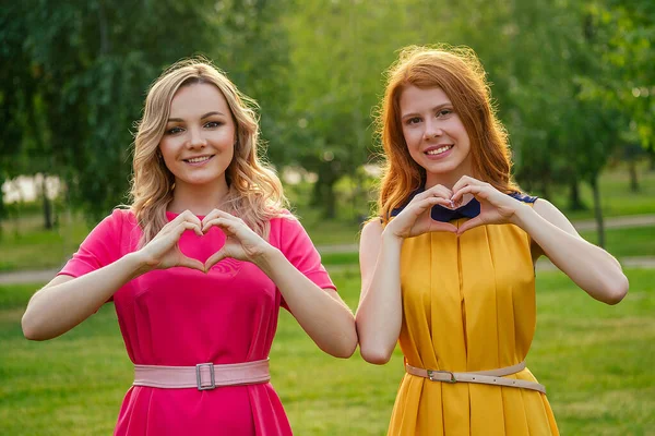 Twee actieve vrolijke vriendinnen mooie jonge gember roodharige Ierse meisje in een gele jurk en europese blonde vrouw in roze jurk handen in de vorm van een hart in het zomerpark — Stockfoto